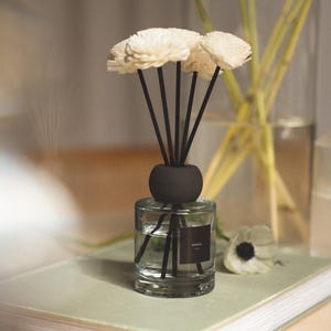 Puede incluir: Un frasco difusor de varillas de vidrio transparente con una tapa esférica negra y varillas negras rematadas con flores de madera de color crema. El frasco contiene un líquido transparente y tiene una pequeña etiqueta cuadrada negra con texto blanco.