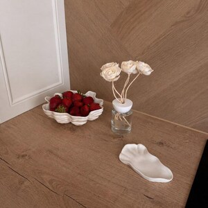 Op de afbeelding: Een witte keramische kom met geschulpte randen gevuld met verse rode aardbeien, een doorzichtige glazen diffuserfles met een witte dop en drie crèmekleurige houten bloemen, en een klein wit keramisch schaaltje met een organische vorm.