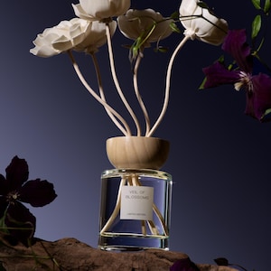 May include: A clear glass reed diffuser bottle with a light wood cap and white flower-shaped reeds. The label reads VEIL OF BLOSSOMS LIMITED EDITION. The diffuser is surrounded by dark purple clematis flowers and green leaves on a piece of wood.
