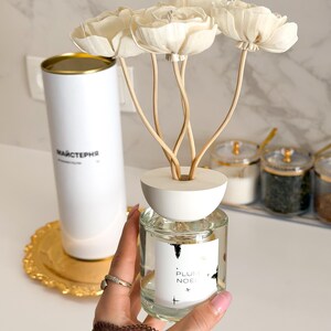 Puede incluir: Un difusor de aromas de vidrio transparente con tapa de madera blanca y tres flores de madera color crema. La etiqueta blanca en la botella presenta el texto PLUM NOEL con pequeños iconos de estrellas. Un tubo cilíndrico blanco se ve al fondo.