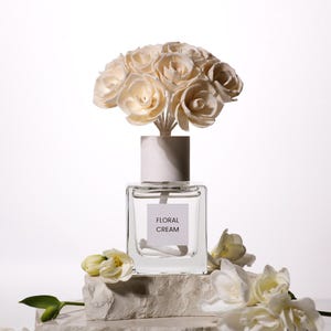 Puede incluir: Un frasco difusor de fragancia de vidrio transparente con tapa blanca y un ramo de flores de madera de color crema. La etiqueta en la parte frontal del frasco cuadrado presenta el texto "FLORAL CREAM" en letras negras. El difusor está rodeado de flores blancas.