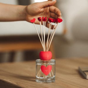 Puede incluir: Un frasco difusor de vidrio transparente con una etiqueta roja en forma de corazón que incluye el texto "ROMANTIC LOVE". El frasco tiene una tapa circular roja y contiene varias varillas de madera finas, cada una con un pequeño corazón rojo.