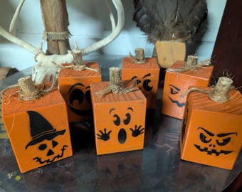 Könnte beinhalten: Sechs orangefarbene Holzblock-Kürbisse mit geschnitzten Jack-o'-Lantern-Gesichtern. Jeder Kürbis hat einen kleinen Holzstiel und Schnurdetails. Ideal für Halloween-Dekorationen.