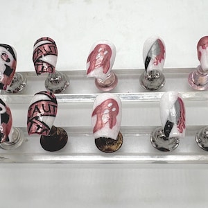 Op de afbeelding: Een set van tien nepnagels met een Halloween-thema. Ontwerpen omvatten het Scream-masker, het woord "AUT", een spook en bloedachtige druppels. De nagels worden gepresenteerd op een heldere, gelaagde standaard.