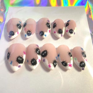 Pode incluir: Um conjunto de dez unhas postiças ovais. Cada unha é pintada com uma base rosa claro, uma ponta branca e um desenho de sprite de fuligem de desenho animado com olhos pretos e estrelas coloridas. As unhas estão dispostas sobre uma superfície branca.