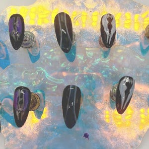 May include: A set of ten artificial nails with various designs. Some nails feature a green and yellow design, others have a black and white pattern, and some display the word "HALO". The nails are displayed on a decorative, iridescent surface.