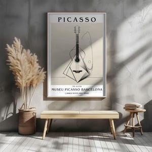 Puede incluir: Un p&oacute;ster enmarcado con una ilustraci&oacute;n en blanco y negro de una guitarra, con el nombre "PICASSO" en la parte superior. Tambi&eacute;n incluye el texto "MUSEU PICASSO BARCELONA". La obra de arte se exhibe en una habitaci&oacute;n con un banco y plantas decorativas.