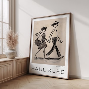 Puede incluir: Impresión artística enmarcada que presenta un boceto en blanco y negro de dos figuras caminando de la mano. La obra de arte, etiquetada como "PAUL KLEE" con "Arte Moderno" debajo, se encuentra sobre un fondo beige. El marco es de color marrón cálido y la impresión se exhibe en una habitación bien iluminada.