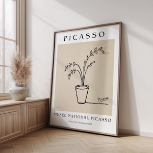 Pode incluir: Uma impressão de arte emoldurada com um desenho minimalista de uma planta num vaso. A impressão tem um fundo bege com o nome "PICASSO" no topo e "MUSÉE NATIONAL PICASSO" na parte inferior. A obra de arte está numa moldura de madeira.