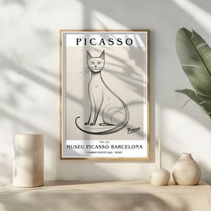 May include: Framed art print featuring a line drawing of a cat, with the name "PICASSO" at the top. The print includes text indicating the artwork is from the Museu Picasso Barcelona. The frame is a light brown color.