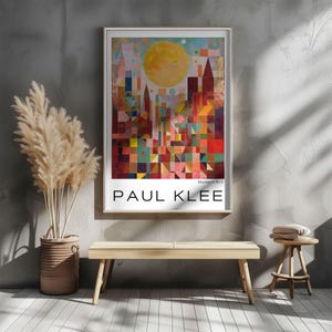 Peut inclure: Une affiche encadr&eacute;e repr&eacute;sentant un paysage urbain abstrait color&eacute; avec un grand soleil jaune. L'affiche est &eacute;tiquet&eacute;e "Modern Art" et "PAUL KLEE" en texte blanc. L'&oelig;uvre est affich&eacute;e sur un mur gris au-dessus d'un banc en bois.