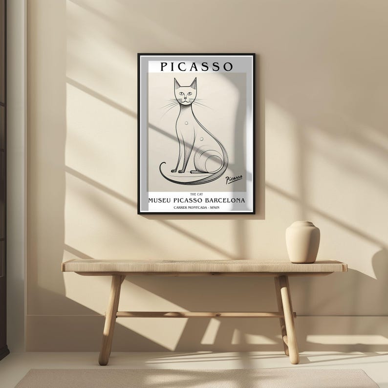 May include: A framed art print featuring a minimalist illustration of a cat, with the name "Picasso" at the top. The print includes the text "Museu Picasso Barcelona" and is displayed above a wooden bench.