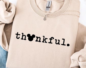 May include: A beige crewneck sweatshirt with the word "thankful" printed in black. The "o" in thankful is replaced with a Mickey Mouse head silhouette. Light blue denim jeans with distressed details are visible.