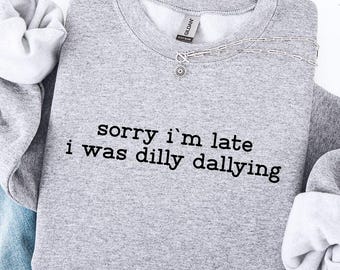 May include: A heather gray sweatshirt with the text "sorry i'm late i was dilly dallying" in black. The sweatshirt is paired with light blue denim jeans with distressed details. A silver necklace is visible.