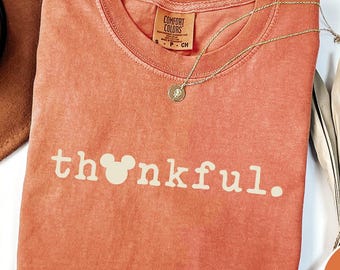 May include: A coral-colored t-shirt with the word "thankful" printed in white, featuring a Mickey Mouse head in place of the "o". A gold necklace and a brown hat are also visible.