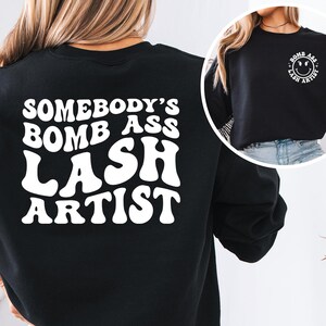 Peut inclure: Sweat-shirt noir avec l'inscription blanche "SOMEBODY'S BOMB ASS LASH ARTIST" au dos. Une petite image montre le devant du sweat-shirt avec un smiley et les mots "BOMB ASS LASH ARTIST".