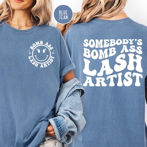 May include: Blue jean-colored t-shirt with white text and a smiley face. The front of the shirt says "BOMB ASS LASH ARTIST" around a smiley face. The back of the shirt says "SOMEBODY'S BOMB ASS LASH ARTIST".