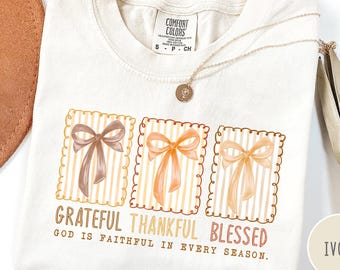 May include: Ivory-colored t-shirt with three bow designs in brown and orange tones. The shirt features the words "Grateful Thankful Blessed" and "God is faithful in every season." A gold necklace and a brown hat are also visible.