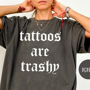 May include: Dark gray oversized t-shirt with the text "tattoos are trashy" in a gothic font. A black circle with the word "pepper" is on the right side of the shirt. The shirt is a casual style.