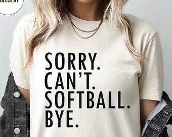 May include: A cream-coloured t-shirt with the text "SORRY. CAN'T. SOFTBALL. BYE." in bold black letters. The shirt is worn with a dark denim jacket. The word "Natural" is in a circle in the upper left corner.