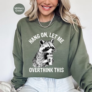 May include: Military green sweatshirt with a raccoon graphic holding a beverage. The text reads "HANG ON, LET ME OVERTHINK THIS" in white. The sweatshirt is a solid color and has a crew neck.