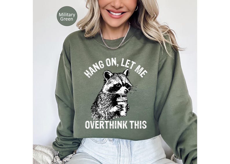 May include: Military green sweatshirt with a raccoon graphic holding a beverage. The text reads "HANG ON, LET ME OVERTHINK THIS" in white. The sweatshirt is a solid color and has a crew neck.