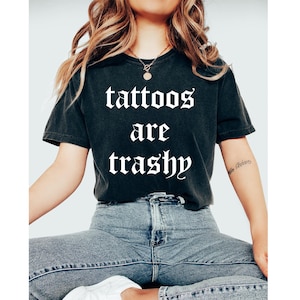 May include: Black t-shirt with the text "tattoos are trashy" in a gothic font. The shirt is worn with blue jeans. The model is wearing a gold necklace.