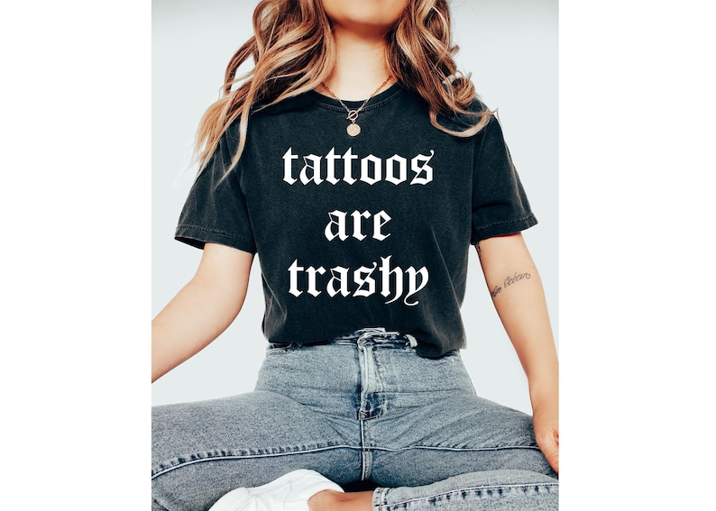 May include: Black t-shirt with the text "tattoos are trashy" in a gothic font. The shirt is worn with blue jeans. The model is wearing a gold necklace.