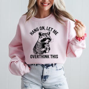 May include: A light pink sweatshirt with the text "HANG ON, LET ME OVERTHINK THIS" above and below a black and white raccoon graphic holding a coffee cup. The sweatshirt is paired with denim shorts.