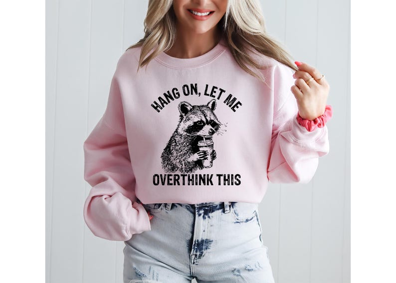 May include: A light pink sweatshirt with the text "HANG ON, LET ME OVERTHINK THIS" above and below a black and white raccoon graphic holding a coffee cup. The sweatshirt is paired with denim shorts.