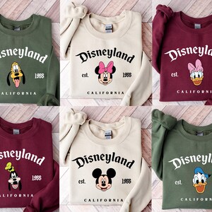 Puede incluir: Sudaderas de cuello redondo en varios colores, cada una con el logotipo de Disneyland y un gr&aacute;fico de personaje. Las sudaderas muestran el texto "Disneyland est. 1955 CALIFORNIA" con diferentes personajes de Disney.