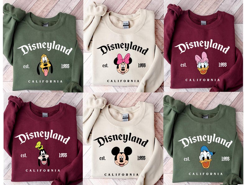 Puede incluir: Sudaderas de cuello redondo en varios colores, cada una con el logotipo de Disneyland y un gr&aacute;fico de personaje. Las sudaderas muestran el texto "Disneyland est. 1955 CALIFORNIA" con diferentes personajes de Disney.