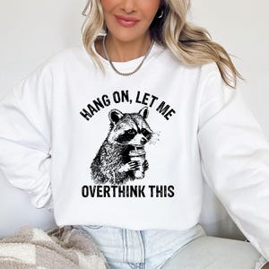 May include: White sweatshirt with a black and white graphic of a raccoon holding a beverage. The text above the raccoon reads "HANG ON, LET ME" and below it reads "OVERTHINK THIS".
