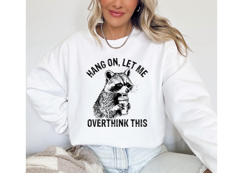 May include: White sweatshirt with a black and white graphic of a raccoon holding a beverage. The text above the raccoon reads "HANG ON, LET ME" and below it reads "OVERTHINK THIS".