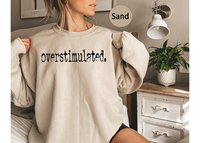 Puede incluir: Sudadera color arena con la palabra "overstimulated" impresa en negro. Esta prenda informal presenta un corte holgado y una fuente sencilla y en negrita. El color neutro la hace vers&aacute;til.