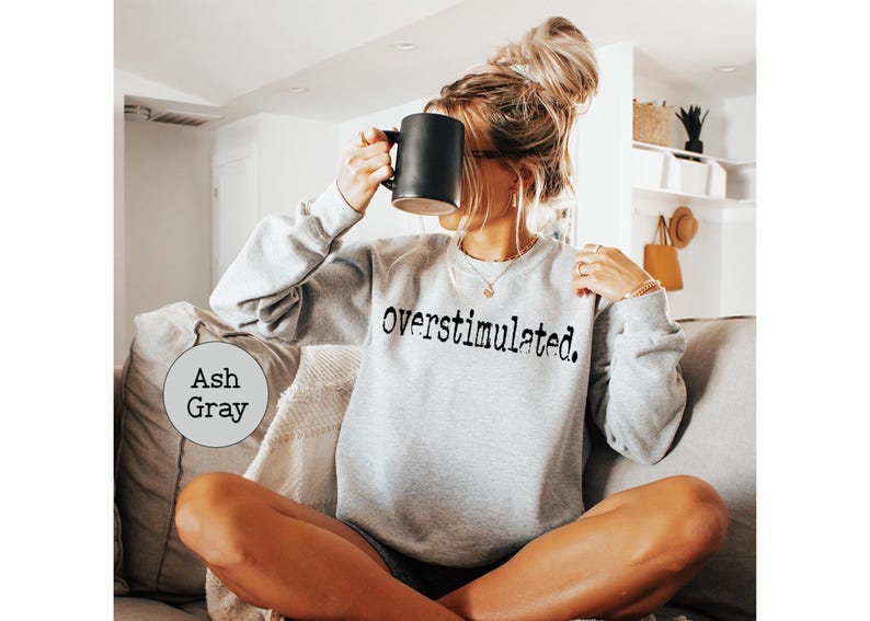 Puede incluir: Sudadera gris ceniza con la palabra "overstimulated" impresa en negro. La persona sostiene una taza negra. La sudadera es de color gris claro. La persona est&aacute; sentada en un sof&aacute;.