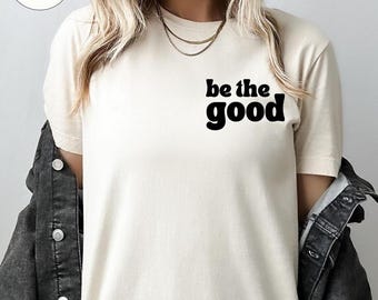 May include: Cream-colored t-shirt with the phrase "be the good" in bold, black letters. The shirt is made of a soft material and is being worn with a black denim jacket. The word "Natural" is in a circle in the upper left corner.