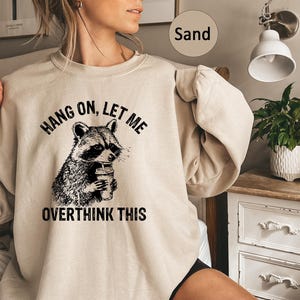 May include: Sand-colored sweatshirt featuring a black and white raccoon graphic holding a coffee cup. The text "HANG ON, LET ME OVERTHINK THIS" is printed above and below the raccoon illustration. The sweatshirt has long sleeves.