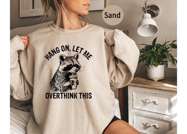 May include: Sand-colored sweatshirt featuring a black and white raccoon graphic holding a coffee cup. The text "HANG ON, LET ME OVERTHINK THIS" is printed above and below the raccoon illustration. The sweatshirt has long sleeves.