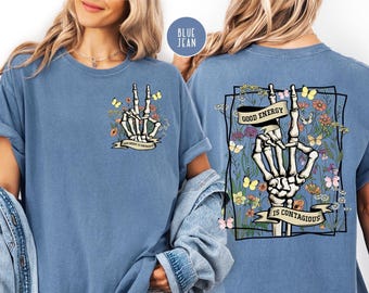 May include: Blue jean-colored t-shirt featuring a skeleton hand in a peace sign gesture, adorned with flowers and butterflies. The front design includes the text "Good Energy Is Contagious" on a banner. The back of the shirt has a similar design.