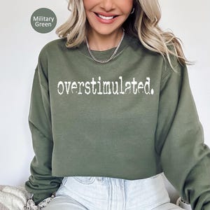 Puede incluir: Sudadera verde militar con la palabra "overstimulated" impresa en blanco. La sudadera es de cuello redondo y tiene mangas largas. El texto est&aacute; en una fuente desgastada.