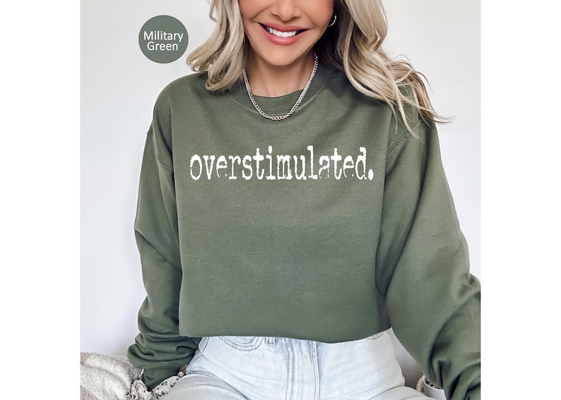 Puede incluir: Sudadera verde militar con la palabra "overstimulated" impresa en blanco. La sudadera es de cuello redondo y tiene mangas largas. El texto est&aacute; en una fuente desgastada.