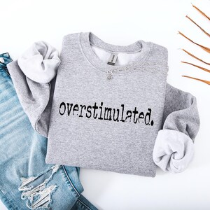 Puede incluir: Una sudadera gris jaspeada con la palabra "overstimulated." impresa en negro. La sudadera se combina con unos vaqueros de mezclilla azul claro con detalles desgastados. Tambi&eacute;n se ve un collar y una hoja de palma seca.
