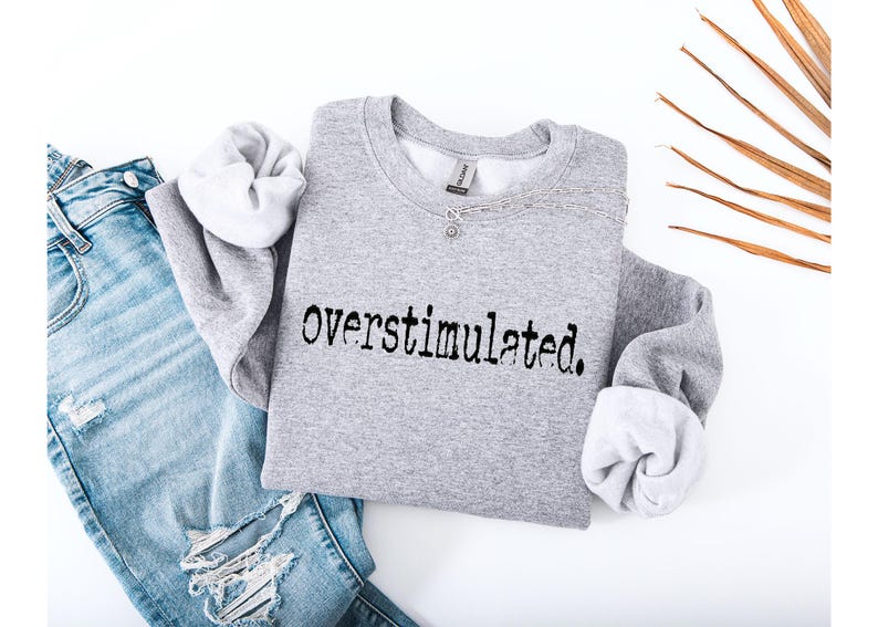 Puede incluir: Una sudadera gris jaspeada con la palabra "overstimulated." impresa en negro. La sudadera se combina con unos vaqueros de mezclilla azul claro con detalles desgastados. Tambi&eacute;n se ve un collar y una hoja de palma seca.