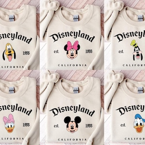 Puede incluir: Sudaderas de color crema con el logotipo de Disneyland y varias caras de personajes de Disney. Las sudaderas incluyen el texto "est. 1955 CALIFORNIA". Los personajes incluyen Mickey Mouse, Minnie Mouse, Goofy, Pluto, Donald Duck y Daisy Duck.