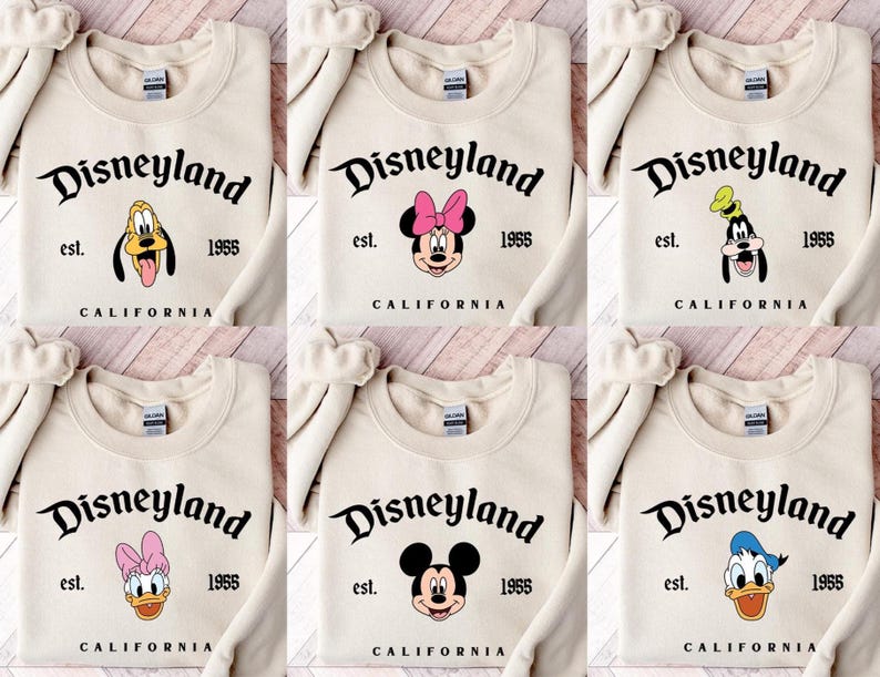 Puede incluir: Sudaderas de color crema con el logotipo de Disneyland y varias caras de personajes de Disney. Las sudaderas incluyen el texto "est. 1955 CALIFORNIA". Los personajes incluyen Mickey Mouse, Minnie Mouse, Goofy, Pluto, Donald Duck y Daisy Duck.