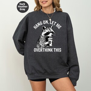 May include: Dark heather gray sweatshirt with a raccoon graphic holding a coffee cup. The text "HANG ON, LET ME OVERTHINK THIS" is printed above and below the raccoon illustration. The sweatshirt is a casual style.