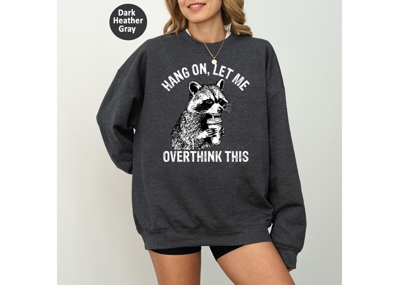 May include: Dark heather gray sweatshirt with a raccoon graphic holding a coffee cup. The text "HANG ON, LET ME OVERTHINK THIS" is printed above and below the raccoon illustration. The sweatshirt is a casual style.