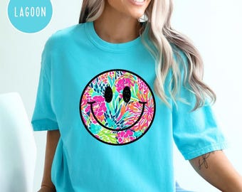 May include: A light blue t-shirt featuring a colorful smiley face design. The smiley face has a black outline and is filled with a floral pattern in shades of pink, green, and orange. The word "LAGOON" is in a light blue circle in the upper left.
