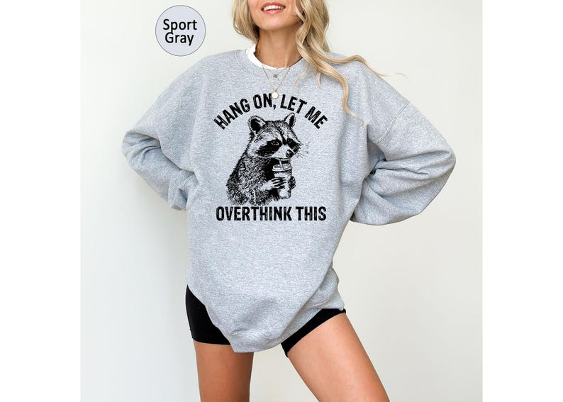 May include: Sport gray sweatshirt with a raccoon graphic holding a coffee cup. The text on the sweatshirt reads "Hang on, let me overthink this". The sweatshirt is a light gray color.
